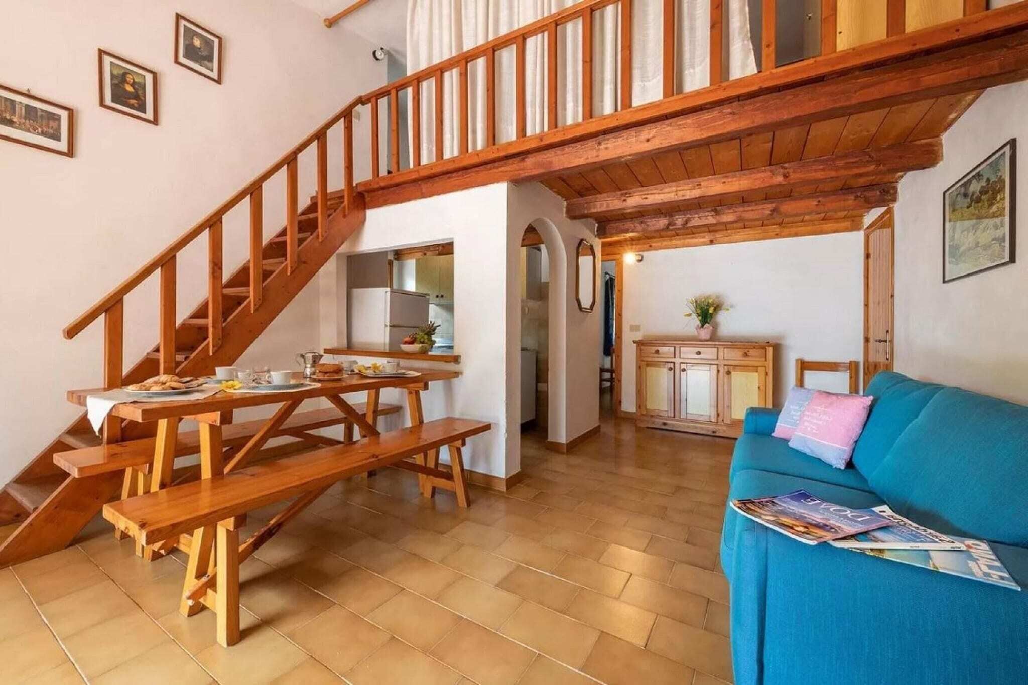 Un'ampia zona giorno con una scala in legno, un tavolo da pranzo con panche, un divano blu e una cucina sullo sfondo.