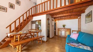 Uma sala de estar ampla com escada de madeira, mesa de jantar com bancos, sofá azul e cozinha ao fundo.