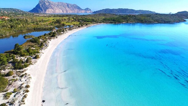 Paisagem costeira com praia, águas azuis cristalinas e montanha ao fundo.