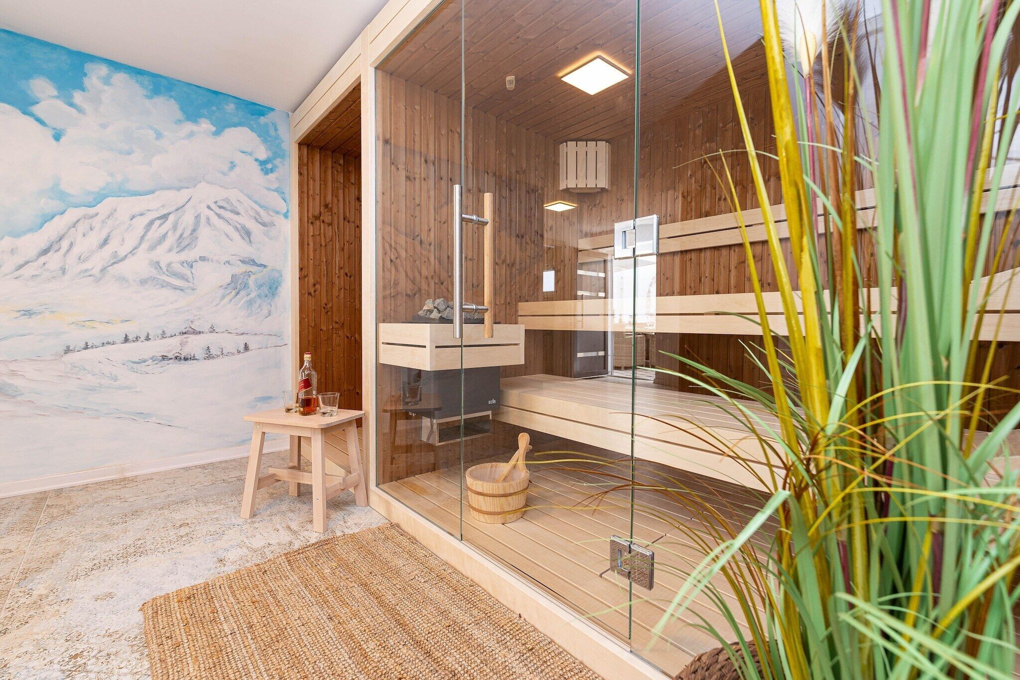 A sauna room with wooden benches, a glass door, and a painting of a snowy mountain landscape.