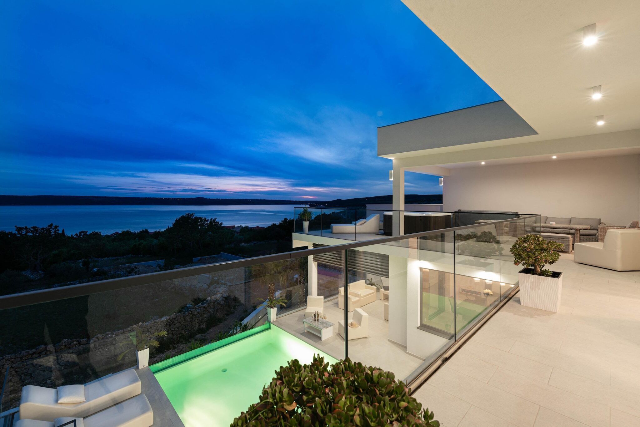 A modern outdoor living space with a glass balustrade, a pool, and a view of the sea.