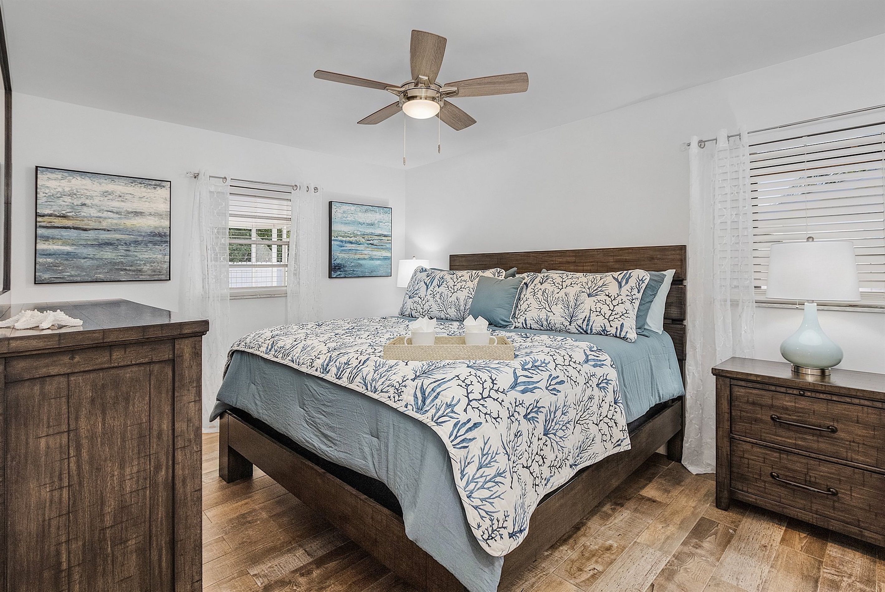 A bedroom with a bed, a nightstand, a bedside table, a ceiling fan, and artwork on the walls.
