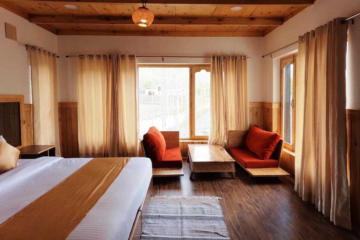 Una habitación con techo de madera, una cama con colcha blanca y marrón, un banco de madera con cojín rojo, una mesita y una ventana con cortinas.