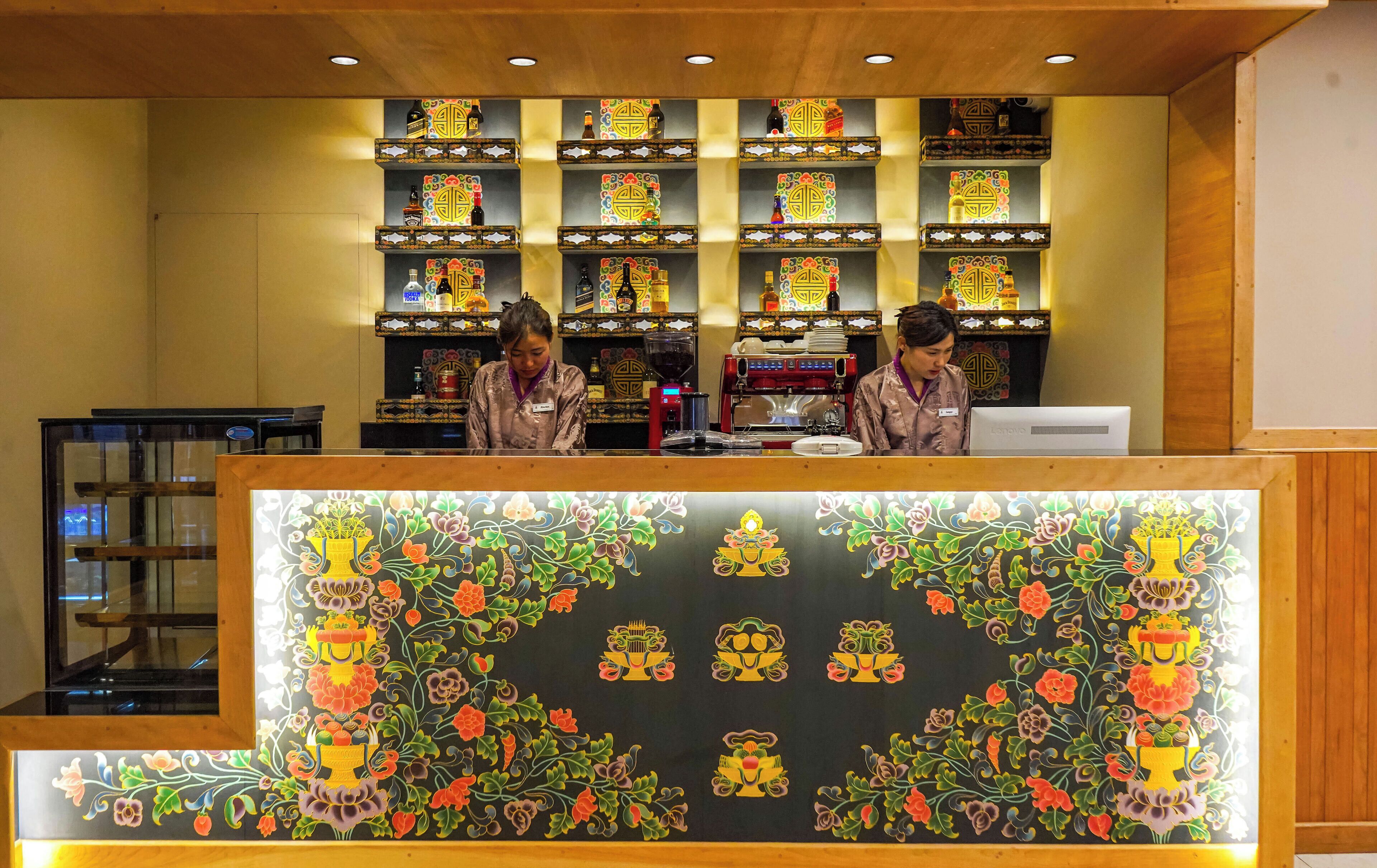 Zona de recepción con mostrador, dos empleados y una pared decorativa con estantes repletos de objetos.