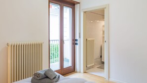 Une chambre à coucher avec un lit, un radiateur, une porte donnant sur un balcon et une salle de bain visible à travers une porte ouverte.