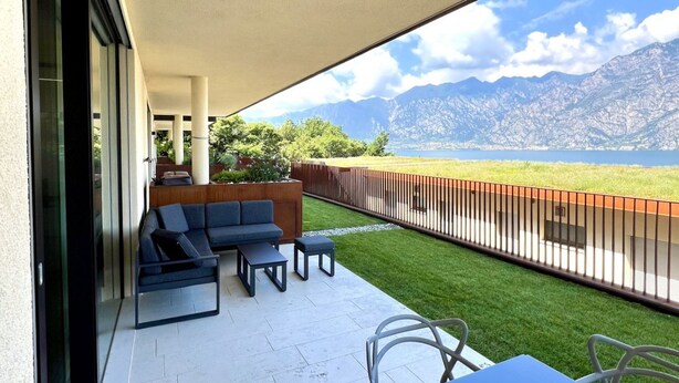 A balcony with a sofa, chairs, and a table, overlooking a grassy area and mountains. - Lakefront Seeblick Le Dimore (Malcesine)