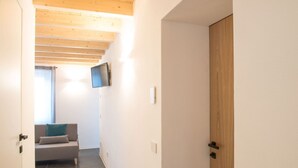 A hallway with a wooden ceiling, a grey sofa with a teal pillow, a television mounted on the wall, and a wooden door.