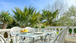 Een terras met een glazen tafel en witte stoelen, omgeven door weelderig groen en palmbomen.