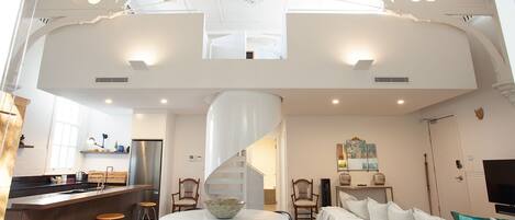 A spacious living area with a high ceiling, a central spiral staircase, and a kitchen area with a refrigerator and bar stools.