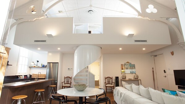A spacious living area with a high ceiling, a central spiral staircase, and a kitchen area with a refrigerator and bar stools.