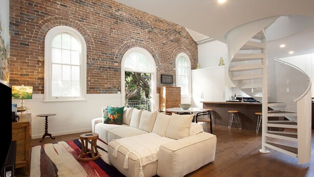 Un salon spacieux avec un canapé blanc, une table basse et une grande fenêtre. On aperçoit un mur en briques et un escalier en colimaçon à l’arrière-plan.