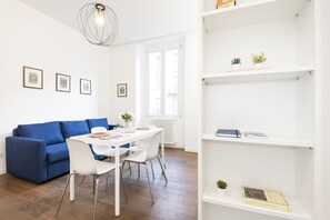 Ein modernes Wohnzimmer mit einem blauen Sofa, einem weißen Esstisch und Stühlen. An der Wand befinden sich gerahmte Bilder, ein Heizkörper und ein Bücherregal mit Büchern und kleinen Pflanzen.