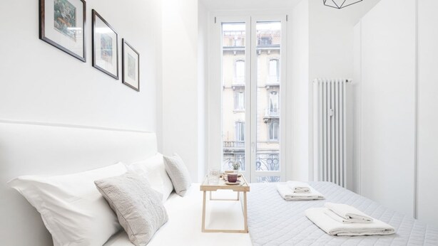 Habitación de hotel minimalista con una cama blanca, una pequeña mesa de madera con una taza y un plato, y vistas a edificios a través de un ventanal grande.