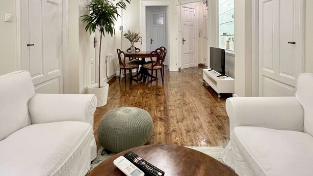 A spacious living area with a wooden floor, white sofas, a dining table with chairs, a television, and a bookshelf. - Magnolia: Spacious neoclassical apartment (Thessaloniki)
