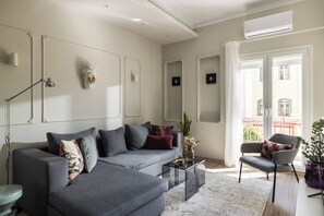 A modern living room with a grey sofa, a side table, and a chair. There is a window with a view of buildings outside. - Liberty by halu! | 3 Bedroom Apartment in Thessaloniki Center (Thessaloniki)