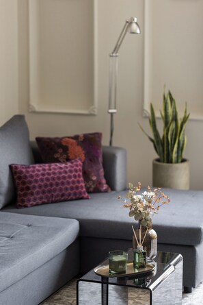 A modern living room with a grey sofa, patterned cushions, a floor lamp, a potted plant, and a glass coffee table with decorative items. - Liberty by halu! | 3 Bedroom Apartment in Thessaloniki Center (Thessaloniki)