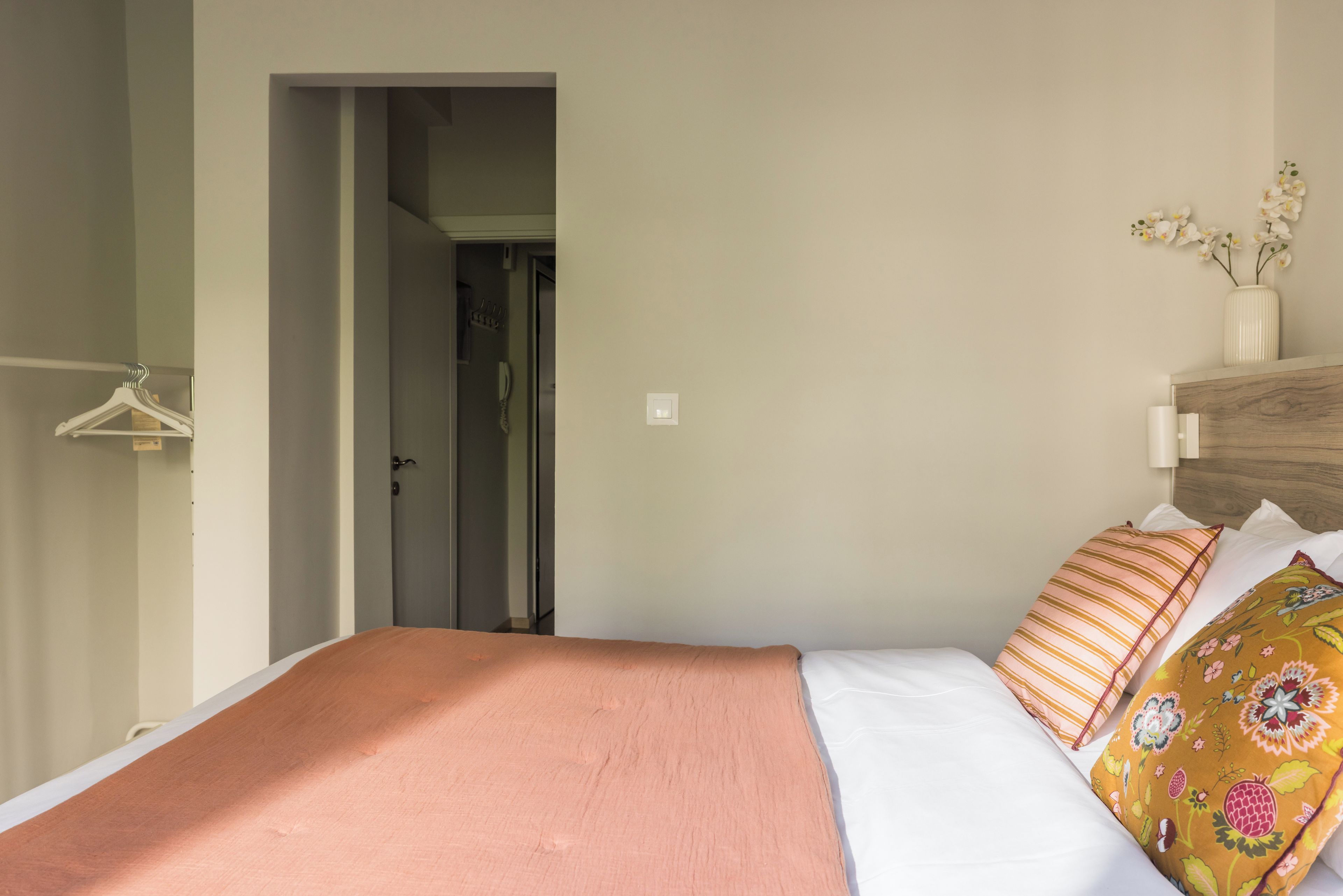 A bedroom with a bed, a bedside table with a lamp, a hanging light fixture, and a door leading to another room.