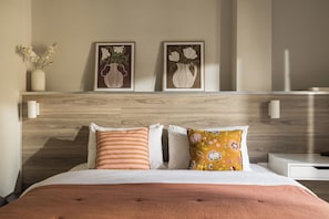 A hotel room with a wooden headboard, two framed floral paintings, a white bedside table, and a bed with striped and floral pillows. - Liberty by halu! | 3 Bedroom Apartment in Thessaloniki Center (Thessaloniki)