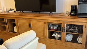 A living room with a large flat-screen TV mounted on the wall, a white sofa, wooden cabinets with dishware, and a coffee maker on the counter. - Bigstone Terrace Suite Tsutsumimachi (Aomori)