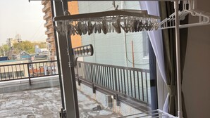 A balcony with a metal drying rack, a window with curtains, and a view of buildings and trees. - Bigstone Terrace Suite Tsutsumimachi (Aomori)