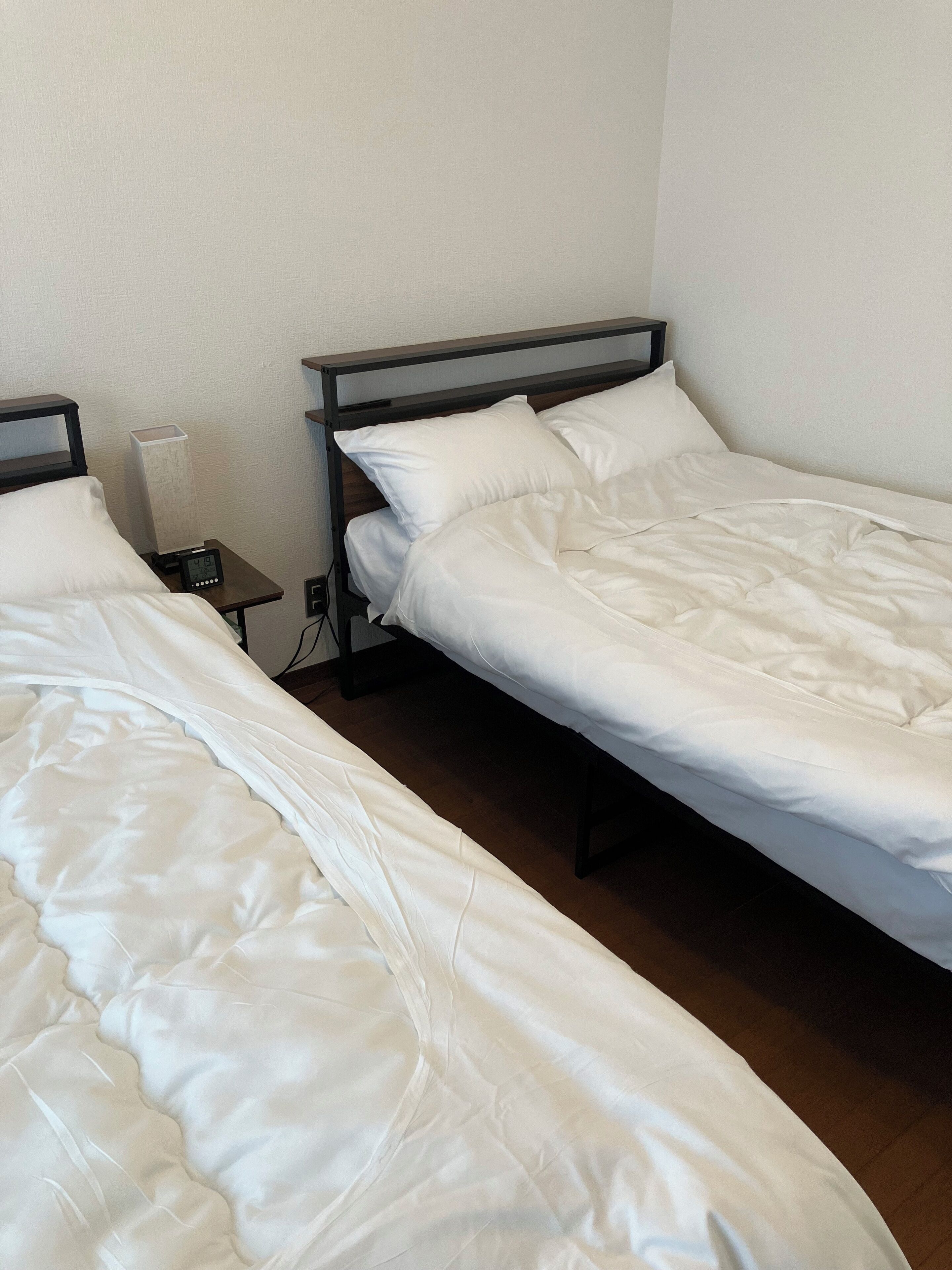 Dos camas individuales con ropa blanca, una mesita de noche pequeña con lámpara y un enchufe de pared.