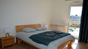 A neatly made bed with a wooden headboard and a view of a parking lot through a large window. - Entre Plage et Ports : Le 49 - Avec Balcon (Courseulles-sur-Mer)