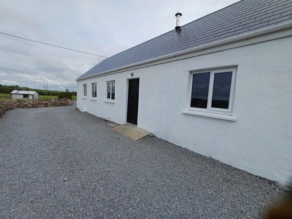 A white building with a grey roof, a gravel driveway, and a stone wall in the background.