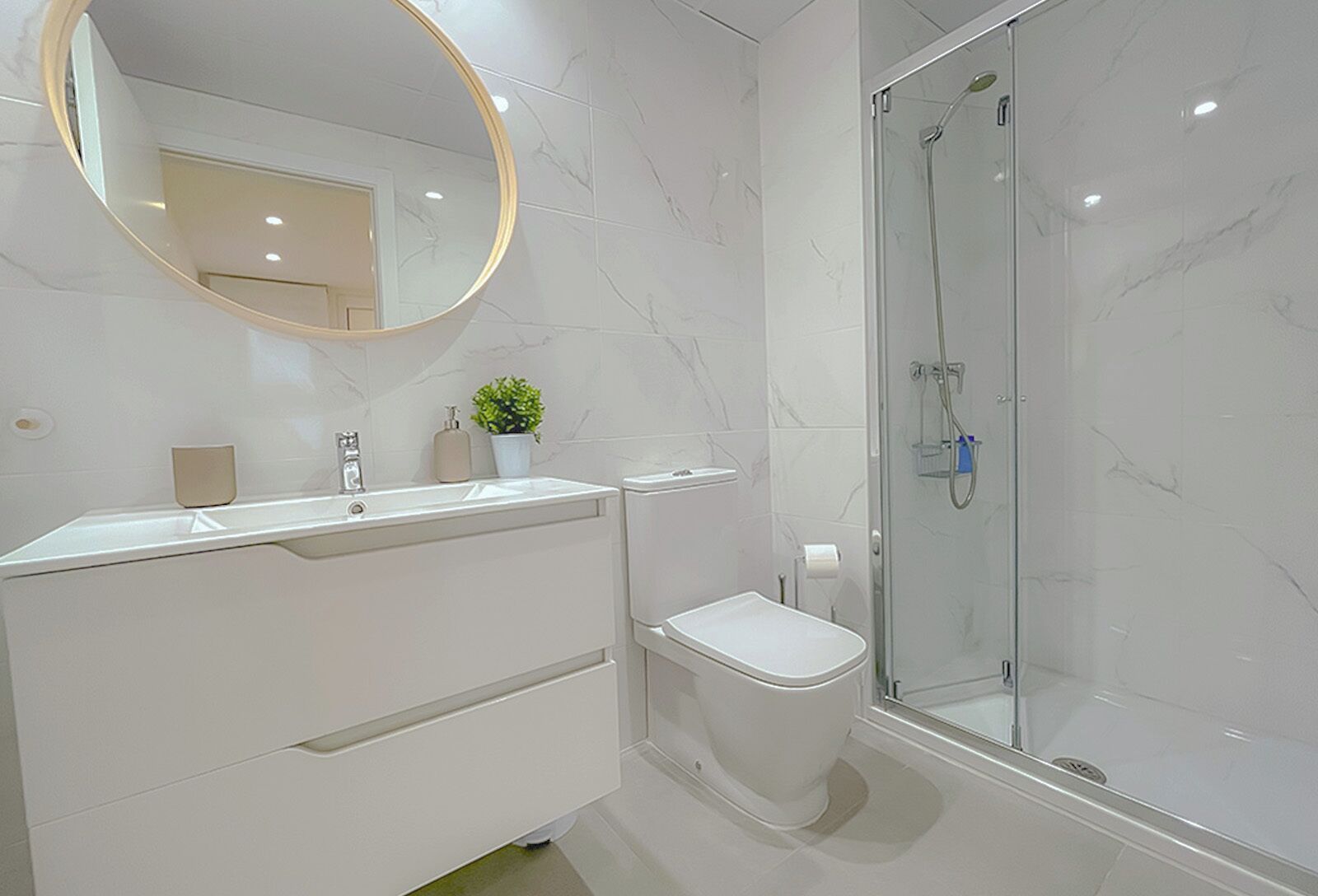Un baño moderno con piso de mármol, tocador blanco con espejo, inodoro y ducha con mampara de vidrio.