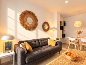 A modern living room with a grey sofa, two round mirrors, a dining area with a white table and chairs, and a basket of oranges on a woven mat. - New apt. at the beach of Daimus - near Gandia (Daimús)