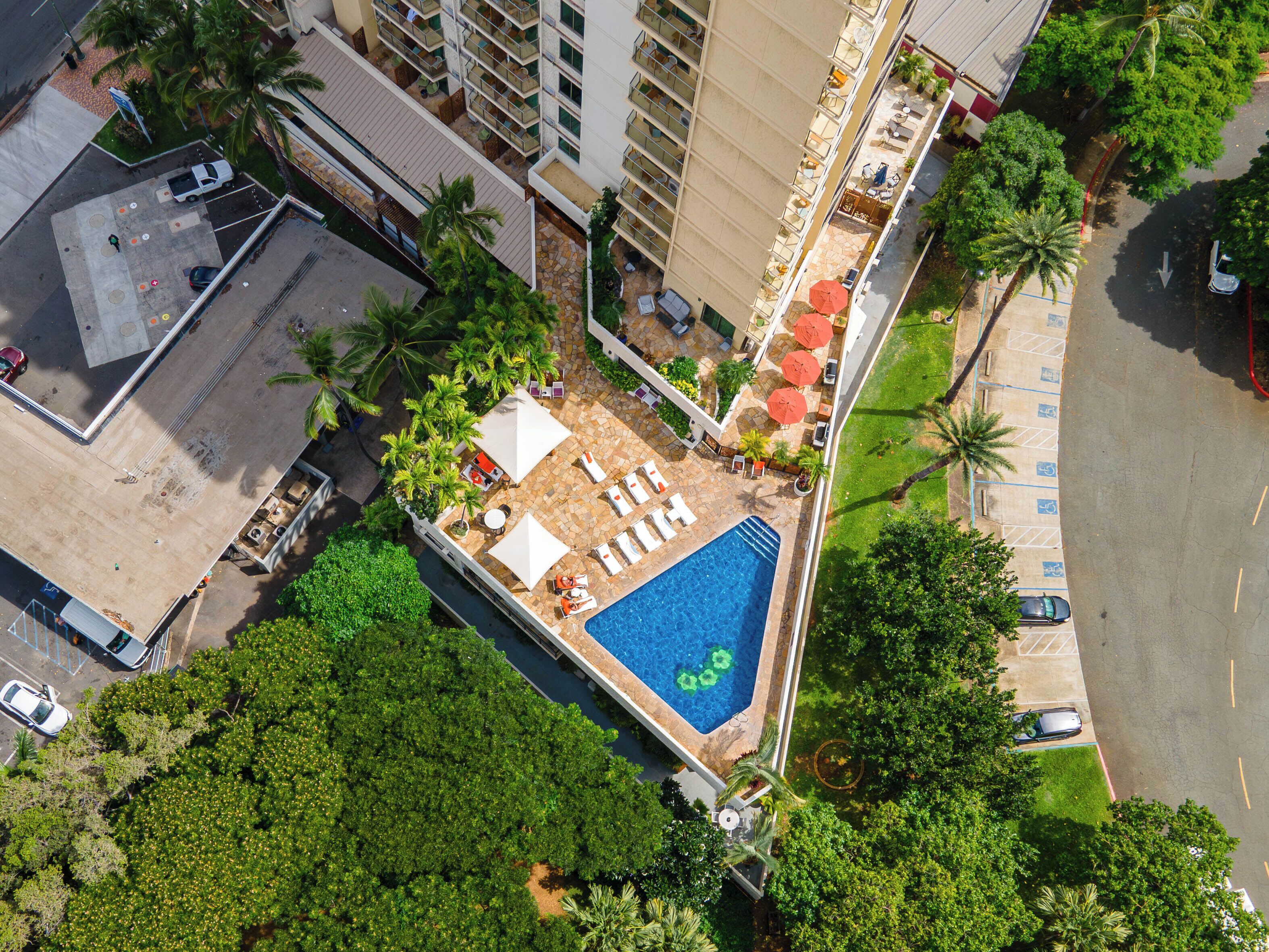 An aerial view of a hotel with a rooftop pool surrounded by lounge chairs and umbrellas, a parking area, and lush greenery.