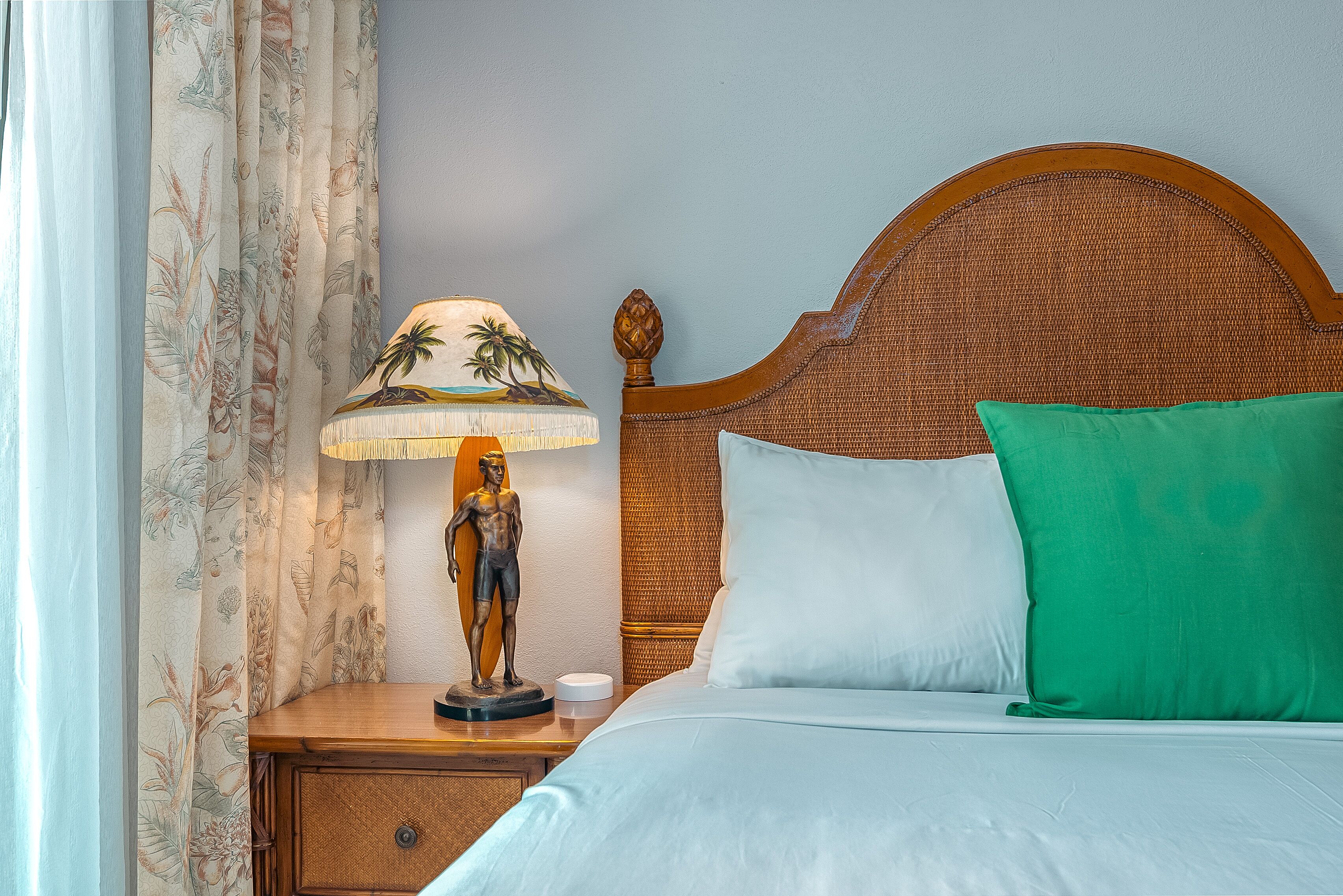 A neatly made bed with white and green pillows, a wooden nightstand with a lamp and a statue, and floral curtains.