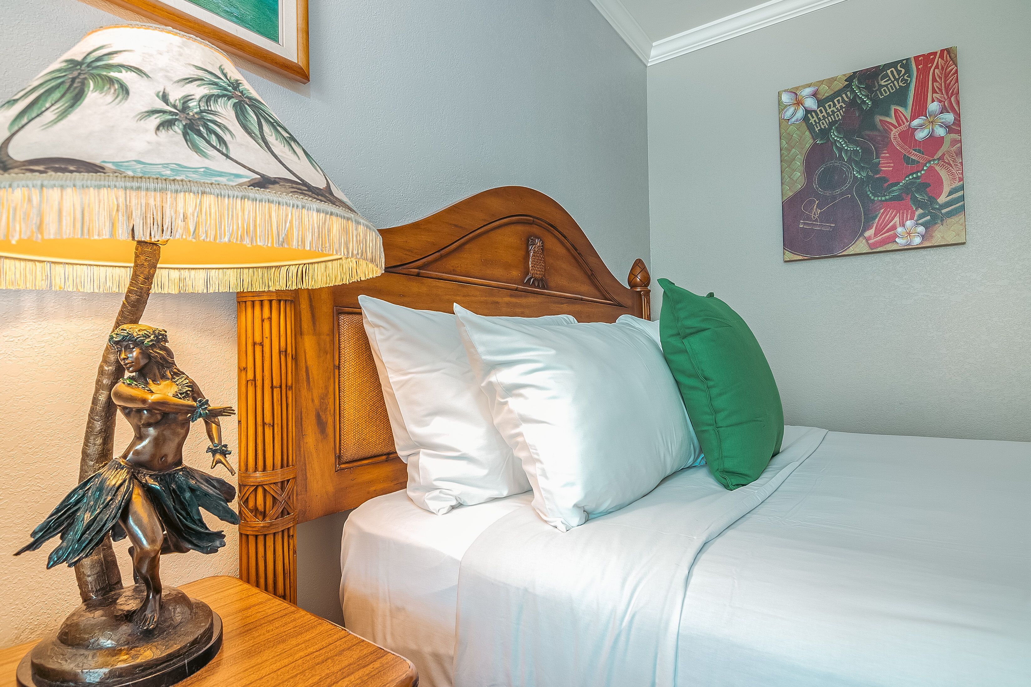 A bed with white and green pillows, a wooden headboard, a wall-mounted painting, and a table lamp with a tropical design.