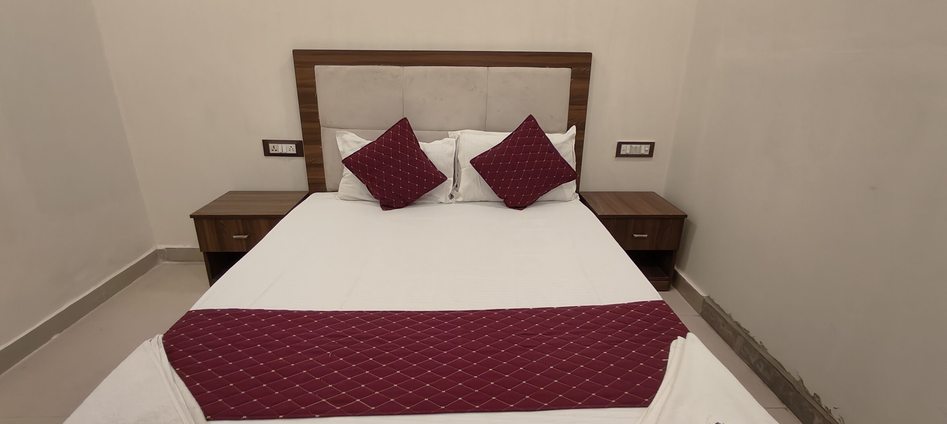 Una cama bien hecha con edredón blanco y dos almohadas color burdeos, flanqueada por mesitas de noche de madera con cajones.