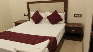 Una cama bien tendida con una colcha blanca y granate, dos almohadas a juego y una mesita de noche de madera con una pequeña mesa al lado.