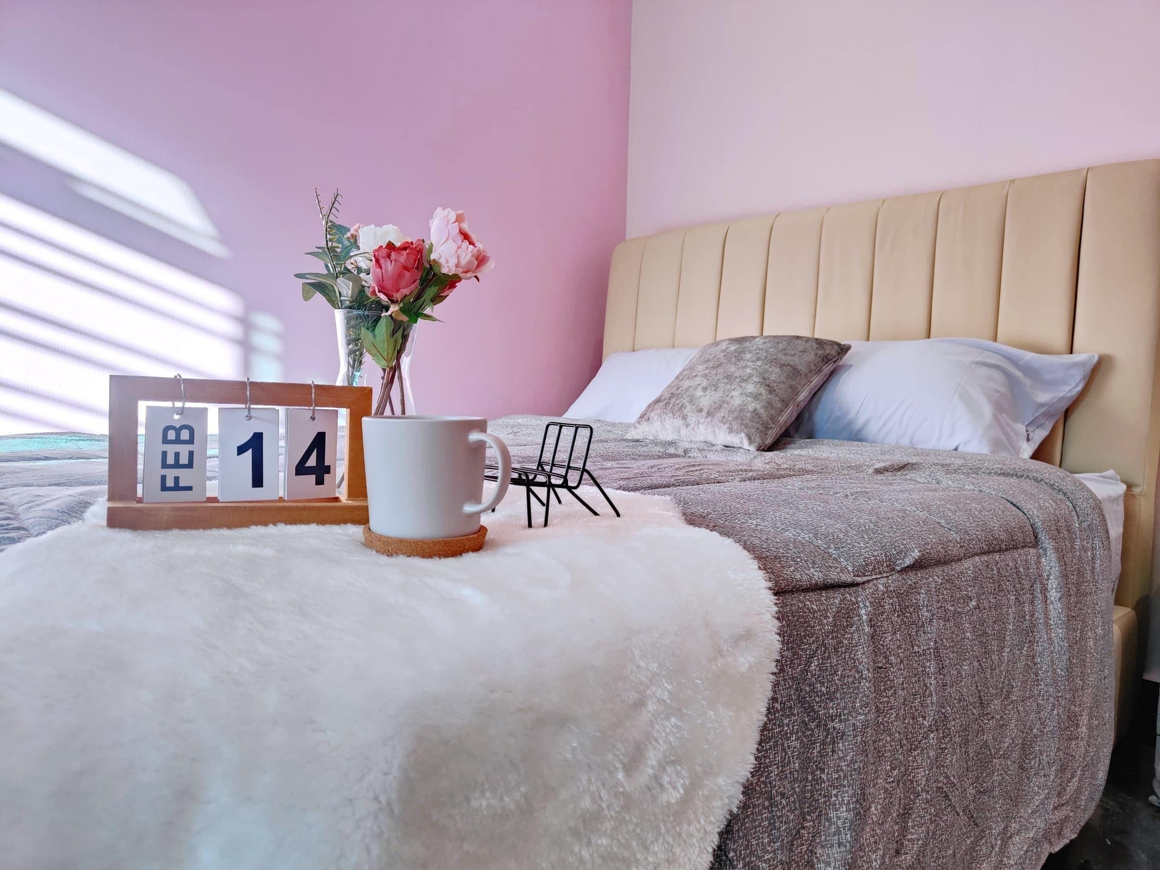 A bed with a beige headboard, a white fluffy pillow, a white mug, a wooden calendar displaying February 14, and a vase with pink flowers.