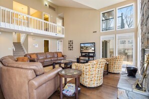 A spacious living room with a brown leather sofa, patterned armchairs, a glass coffee table, a television, and a stone fireplace.