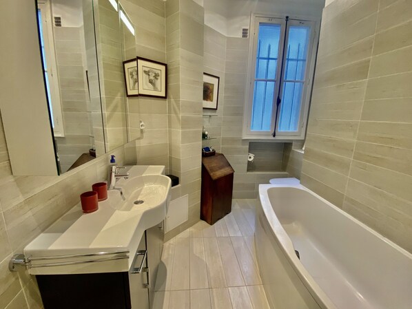 A modern bathroom with a white sink, mirror, and bathtub. There is a window with blinds, a small shelf with toiletries, and a framed picture on the wall. - BEL Appartement Haussmanien, 125 M2, 4 Pieces, 2 CH, 2 Sdb, Proche Gare DE Lyon (Paris)