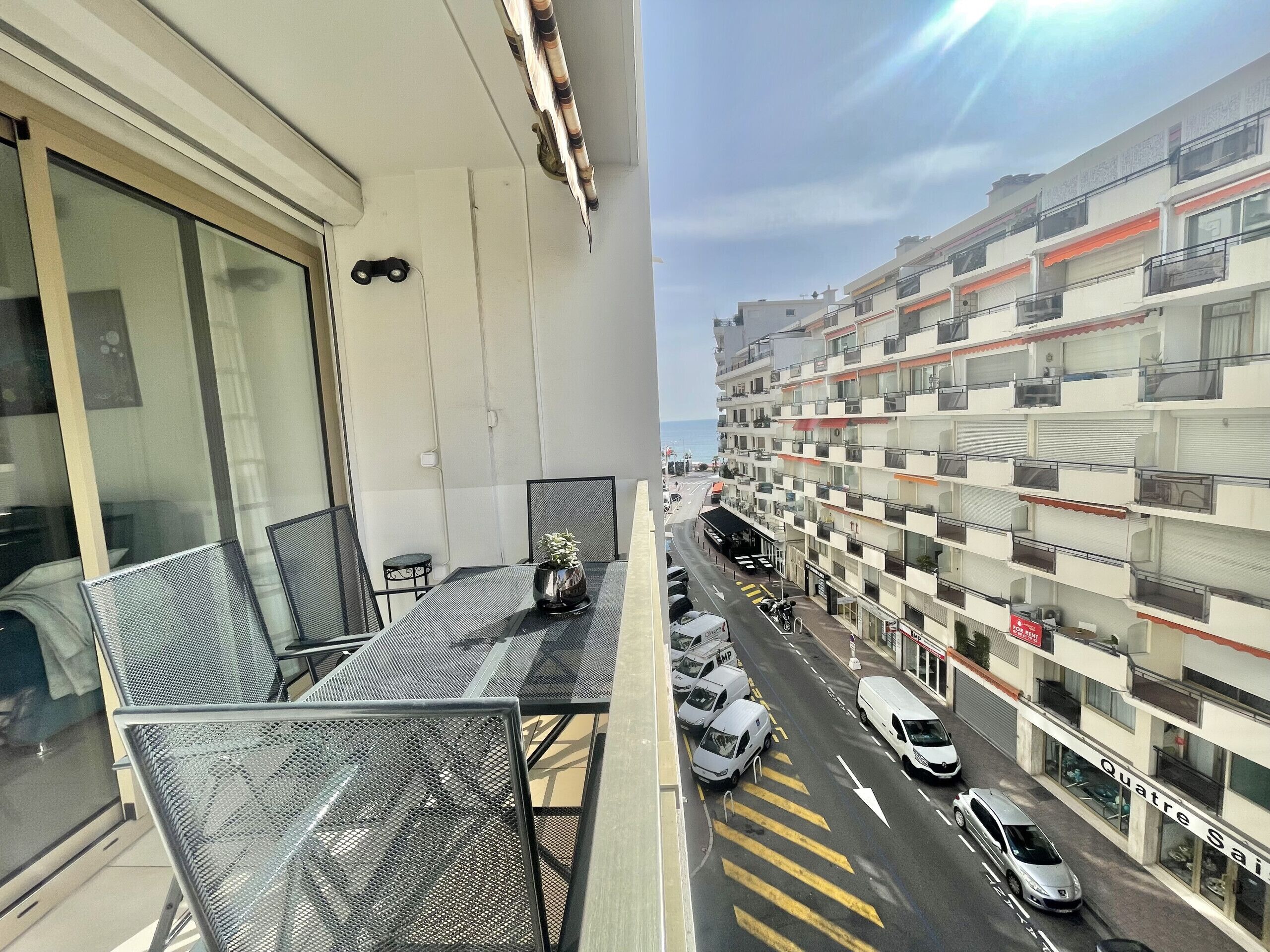 Ein Balkon mit Metallstühlen und einem Tisch, der auf eine Straße mit parkenden Autos und ein mehrstöckiges Gebäude mit Balkonen blickt.