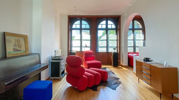 Ein Zimmer mit Holzboden, einem Klavier, roten Sitzmöbeln, einem Schrank und großen Fenstern.