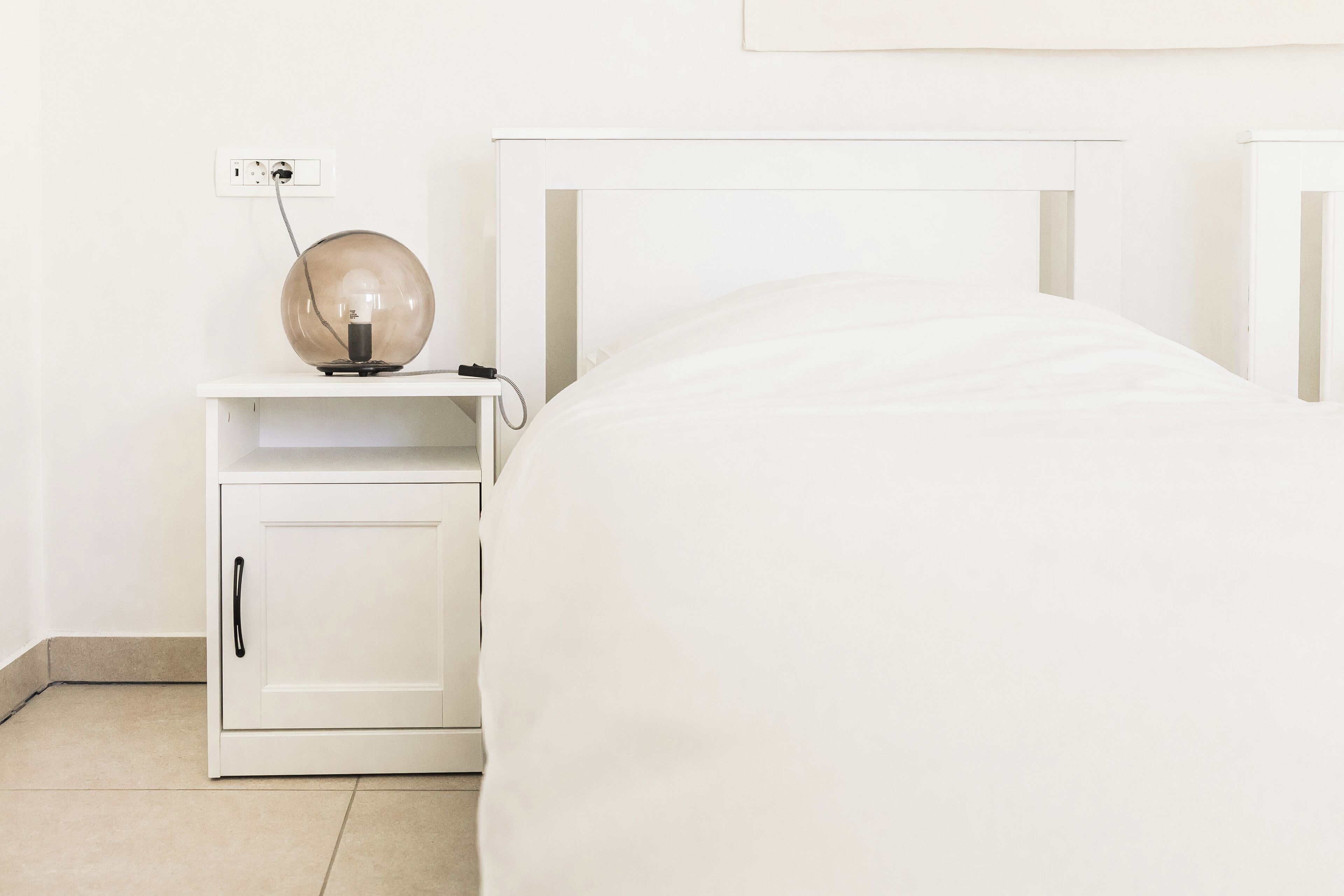 Une table de chevet blanche avec une lampe, un lit blanc et une commode blanche.