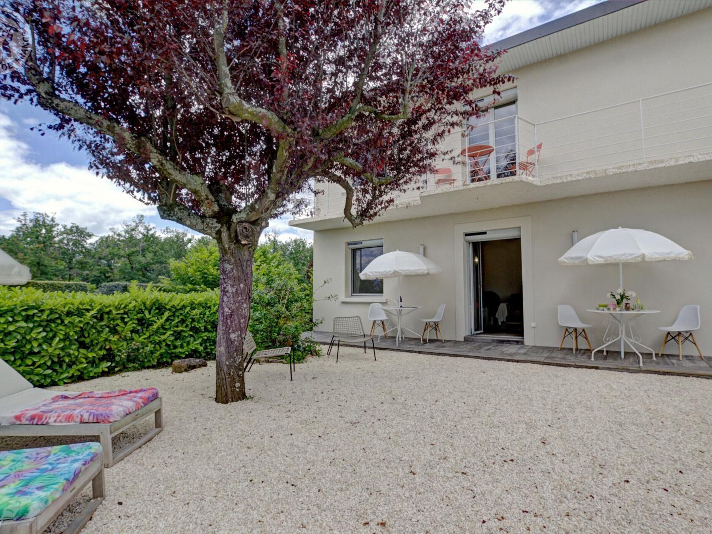 A building with a white exterior, a tree with red leaves, outdoor seating with white umbrellas, and a gravel patio.