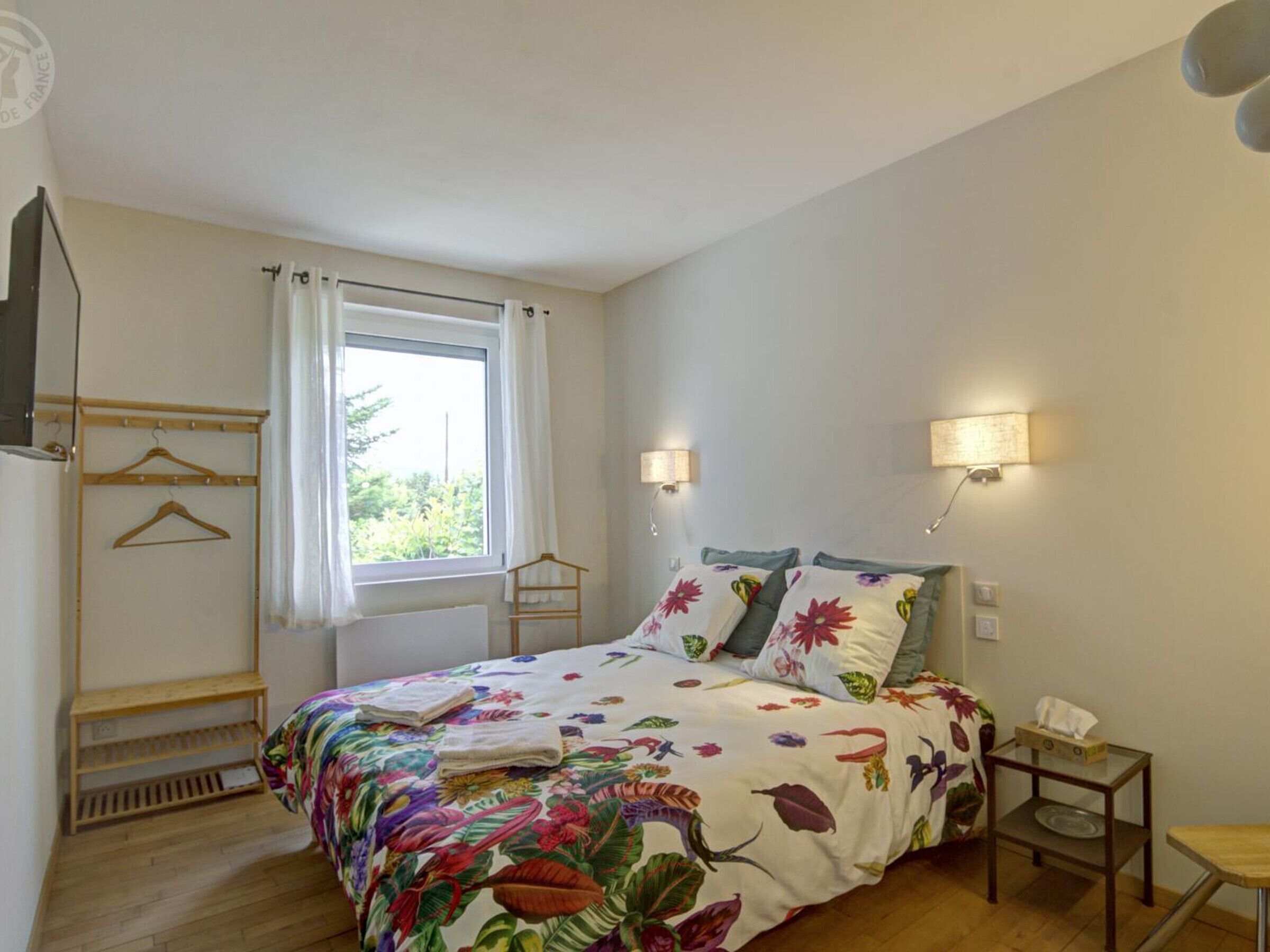 A bedroom with a bed, a window with curtains, a bedside table with a lamp, and a small wooden shelf.