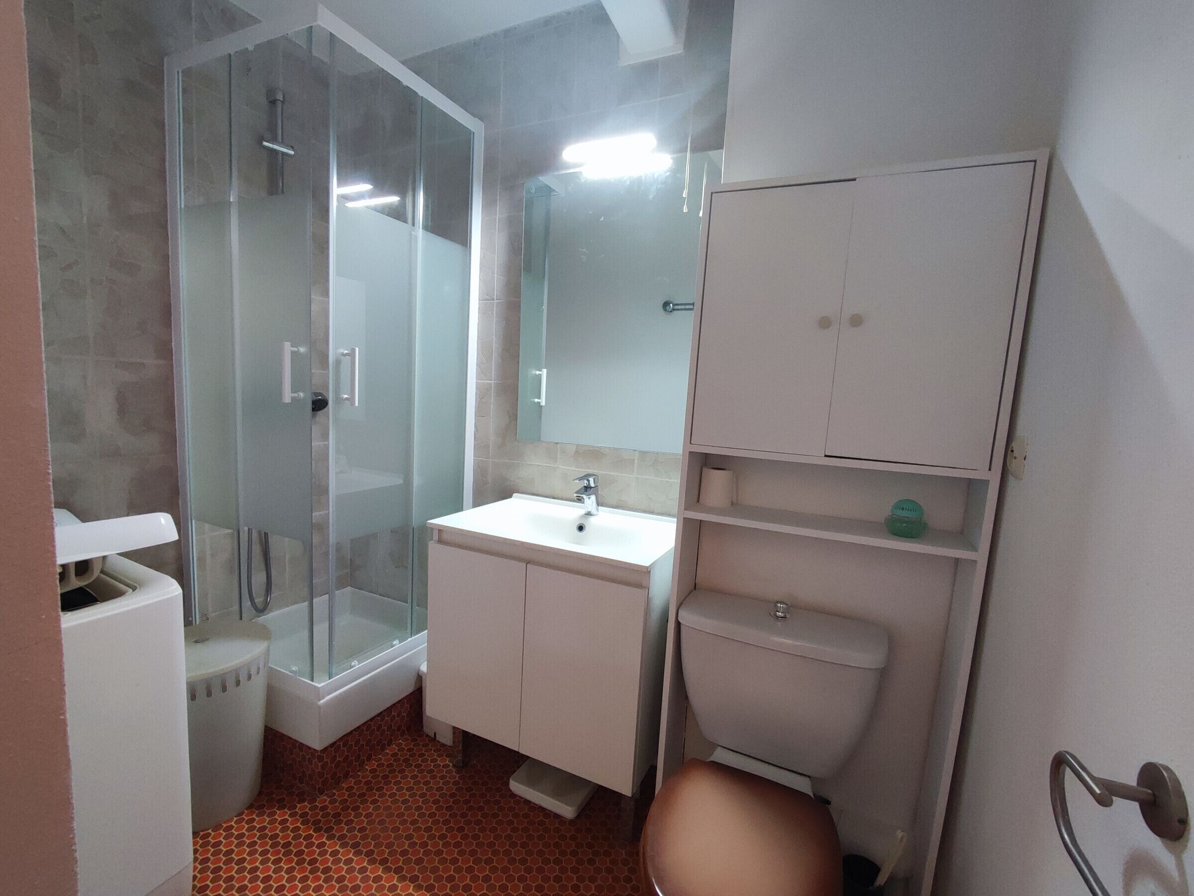 A bathroom with a glass-enclosed shower, a white sink, a toilet, and a cabinet with shelves.