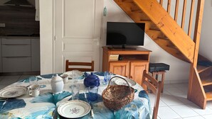 A dining area with a table set for two, a television on a wooden stand, a staircase, and a kitchen in the background.