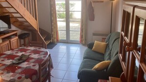 A living room with a wooden staircase, a sofa, a table with a patterned tablecloth, and a view through a glass door to the outside.