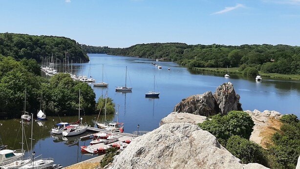 A river with boats docked on both sides, surrounded by greenery and rocky outcrops.