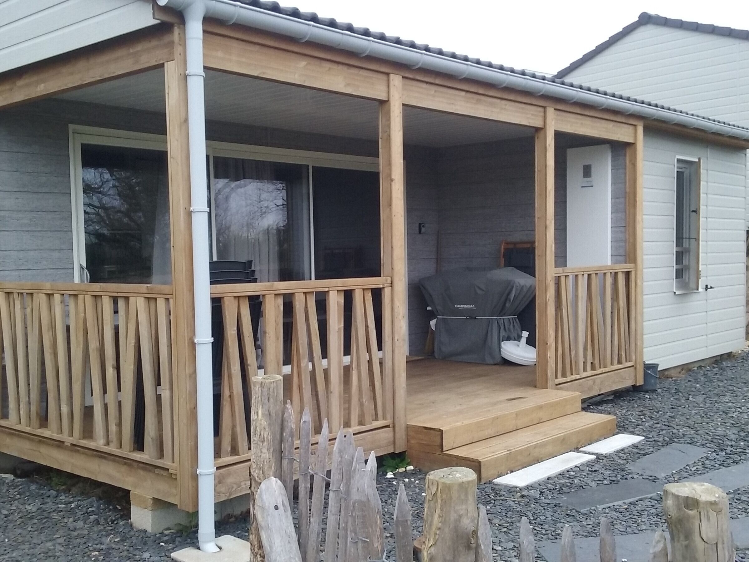 Une maison de plain-pied avec un porche en bois, un espace extérieur couvert aménagé en salon et une fenêtre bien visible.