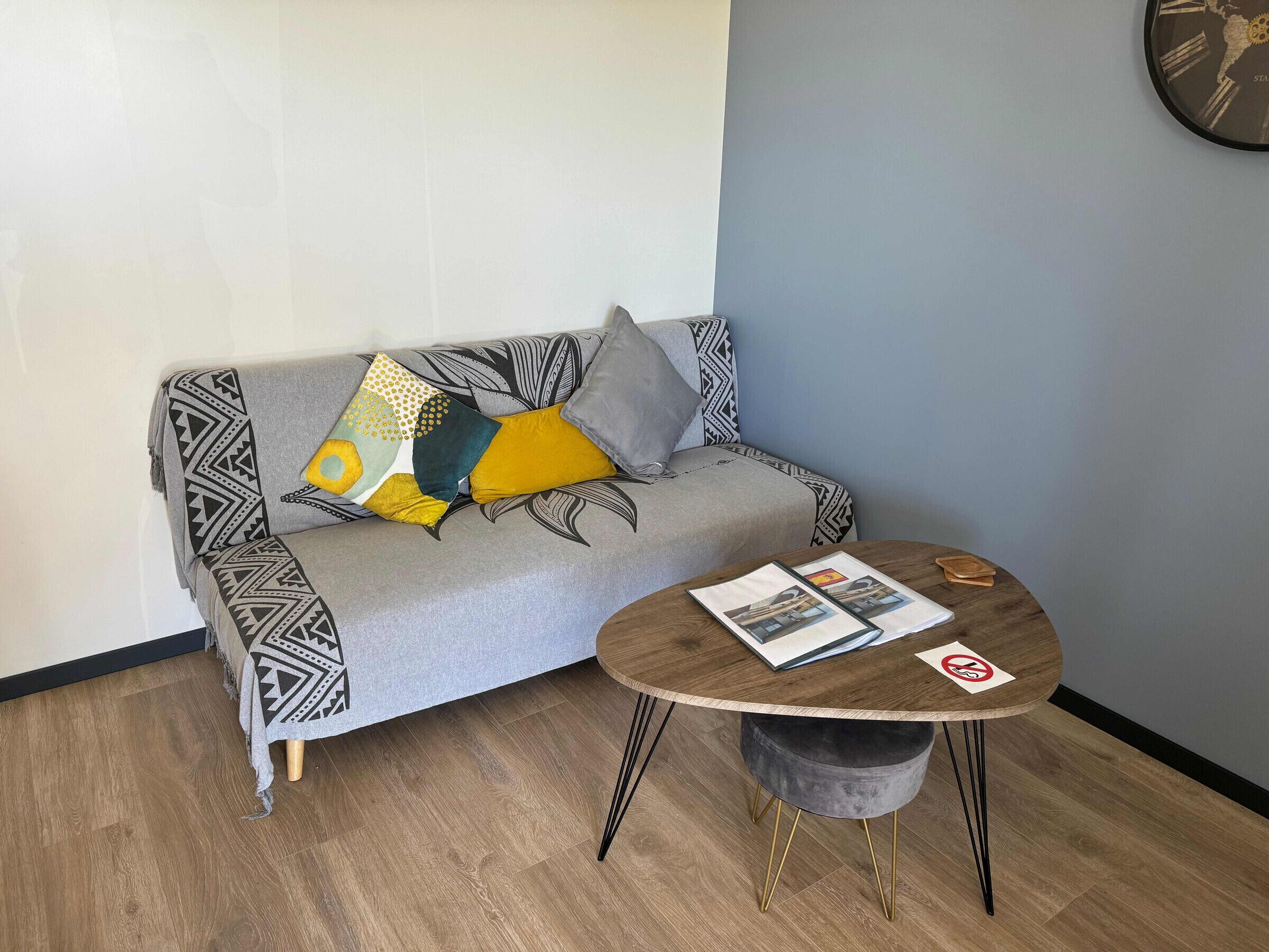 Un salon moderne avec un canapé gris agrémenté de coussins à motifs, une table basse ronde en bois sur laquelle sont posés des magazines, et un petit pouf gris.