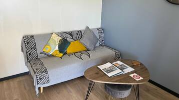 Un salon moderne avec un canapé gris agrémenté de coussins à motifs, une table basse ronde en bois sur laquelle sont posés des magazines, et un petit pouf gris.