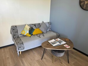 A modern living room with a grey sofa adorned with patterned cushions, a round wooden coffee table with magazines, and a small grey ottoman.